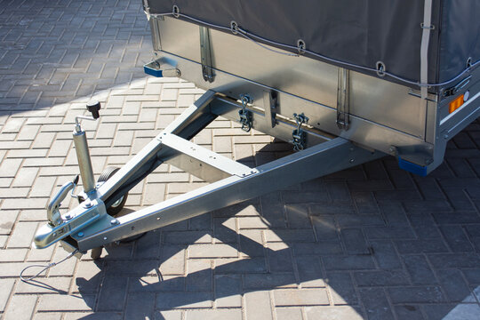 Galvanized steel utility trailer with a grey tarp cover parked on pavement. Towing hitch mechanism, safety cable, and adjustable jockey wheel, representing transportation and logistics.