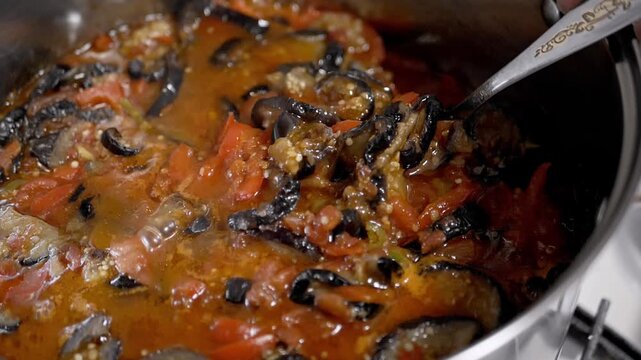 A silver spoon stirs a steaming vegetable stew with eggplant and tomatoes in a metal pot. The close-up view highlights the texture of the cooked vegetables during preparation.