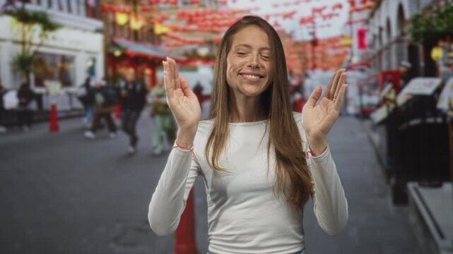 Woman smiling with hands raised palms facing forward wearing white shirt and red string bracelets on a busy street lined with lanterns and shops; joy curiosity.