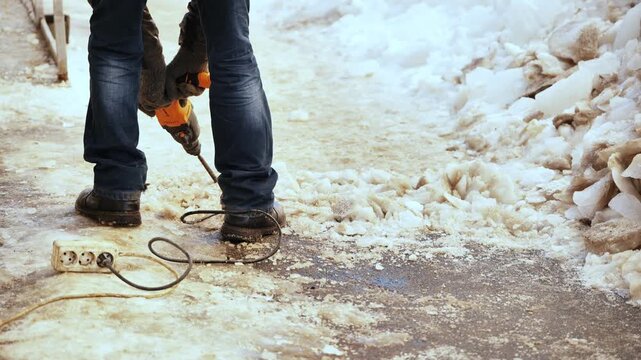 Worker using jackhammer to remove ice from a snow-covered sidewalk during winter