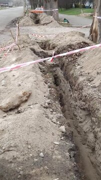 Deep earthen trench dug for utility pipes or cables in an urban park area. Red and white warning tape marks the hazardous construction zone.