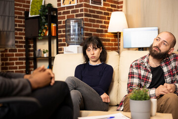 Fototapeta premium Caucasian woman with sad expression sits on couch during therapy session, looking worried at camera beside boyfriend. Young couple listening to african american therapist in private clinic.