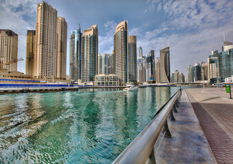 Obraz premium Scenic waterfront view of Dubai Marina skyline with luxury skyscrapers, turquoise water, and a modern promenade walkway under cloudy skies.