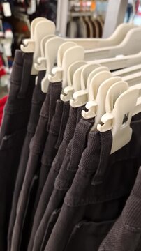 Row of dark-colored pants hangs on white plastic clip hangers on a retail clothing rack. Camera moves slowly along the line of trousers, showing a blurred store background. Background, texture.