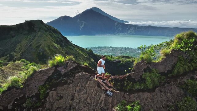 Caucasian man climbing dusty mountain ridge with Lake Batur and volcanic peaks in background, enjoying outdoor sports adventure and explore active volcano landscape on Bali island. Aerial drone flight
