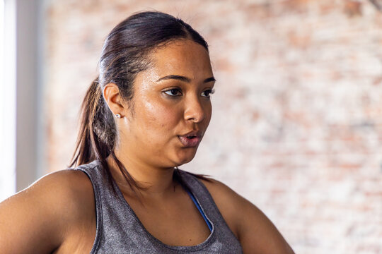 Adult female breathing in fitness studio wearing gray tank, blue bra strap, ponytail, stud earrings