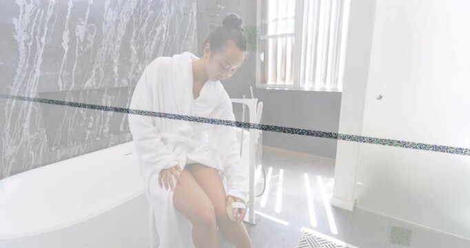 Mid-adult woman in bathrobe seated on tub edge, looking down, applying skincare, moisturizing shin