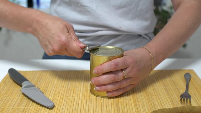 A person uses a can opener to open a canned food item on a bamboo mat in a kitchen. A knife and a fork are on the table.