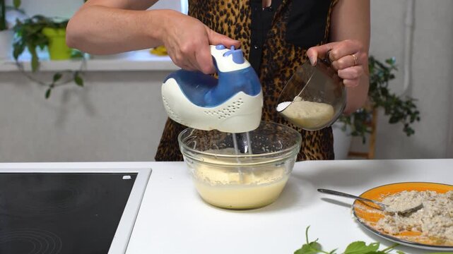 A person uses an electric mixer to combine ingredients in a bowl. There are other items on the kitchen counter.