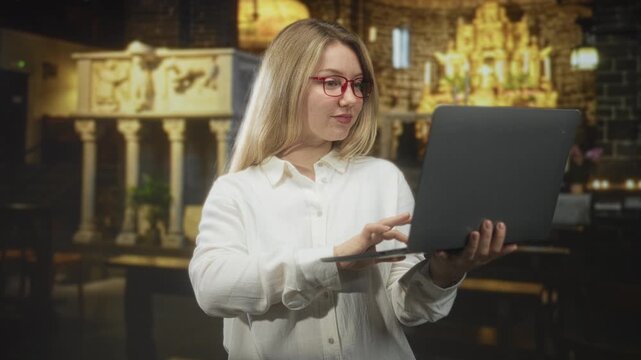 Woman holding laptop and typing with one hand while supporting it with the other in an interior building showing an altar and stone columns; focus productivity.