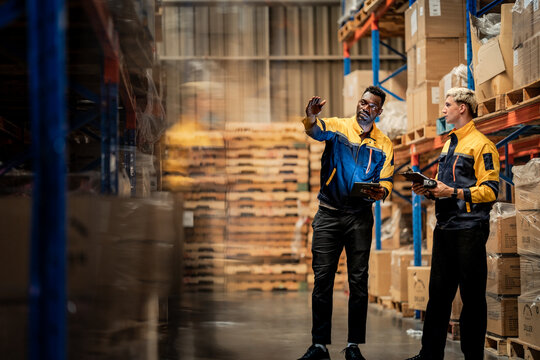 African American and Caucasian logistics staff looking up at cargo rack. Industrial teamwork managing modern supply chain distribution in storehouse.