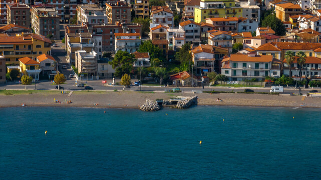 Aerial view on the seafront of Sapri, Campania, Italy. It's a Mediterranean coastal town featuring terracotta-roofed buildings alongside a beach. Small rock groynes extend into the clear blue sea.
