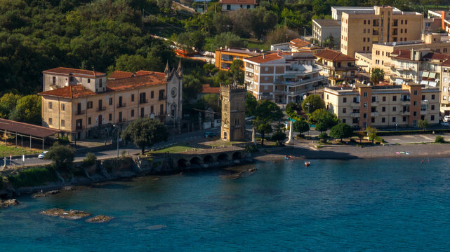 Aerial view on the seafront of Sapri, Campania, Italy. It's a Mediterranean coastal town featuring terracotta-roofed buildings alongside a beach. Small rock groynes extend into the clear blue sea.