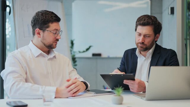 Two professional businessmen engaged in focused discussion, reviewing documents at office desk. Interaction conveys serious planning and collaboration for project success. Concentrated, confident.