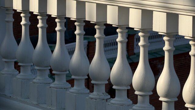 Row of white classic wooden balusters on an outdoor balcony illuminated by sunlight with deep shadows, brick building in background.