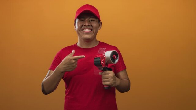 Man wearing red cap and red shirt holding packing tape dispenser and points finger to tape dispenser in studio orange background; friendly service.