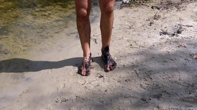 Man's feet covered in thick black mud stand on a sandy beach by the water's edge, an unsettling image of pollution.