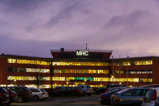 Lund, Sweden - November 19 2018: Illuminated commercial corporate office building with MHC sign on roof and a full car parking lot against a dramatic purple twilight sky