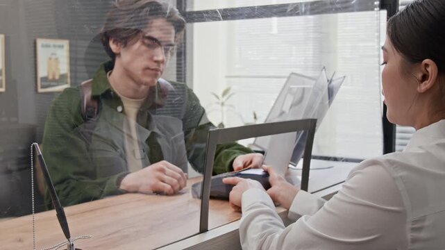 Bank teller holding terminal while young man paying with credit card and dialing pin in modern bank office