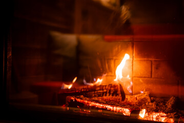 Naklejka premium Charred logs and embers glow in a brick lined fireplace at a San Domenico lodge in the Italian Alps, with warm wood interiors and soft cushions visible beyond.