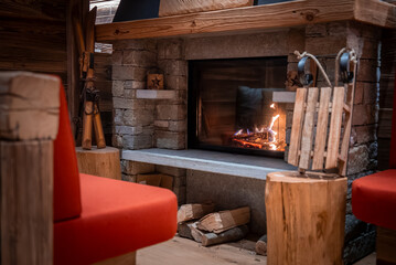Naklejka premium A rustic stone and timber fireplace glows behind glass, split logs sit on the hearth, red benches and wood stools frame vintage skis and a sled in San Domenico.