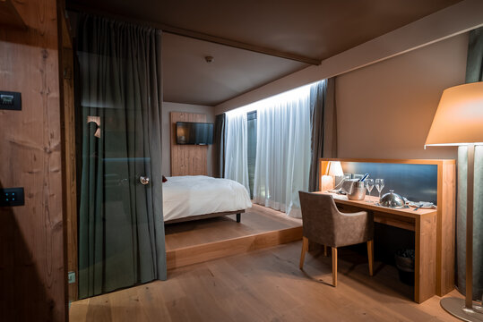 Split level Alpine room in San Domenico, Italian Alps, shows warm wood, soft ambient lighting, bed facing wall TV, desk with lamp and glassware, muted earth tones.