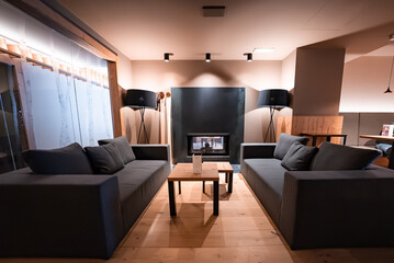 Naklejka premium Two charcoal gray sofas face wooden tables by a built in fireplace in San Domenico, Italian Alps. Tripod lamps and recessed lights warm the rustic wood interior.