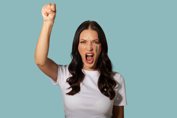 Young woman with Italian flag painted on her cheek raising fist and shouting, standing over blue...
