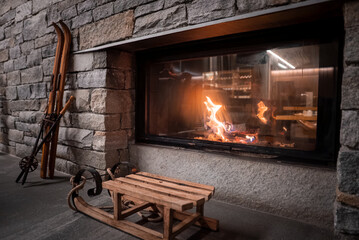 Naklejka premium A modern glass front fireplace set in rough stone casts warm light. A vintage sled and classic wooden skis lean nearby in a San Domenico Italian Alps lodge.