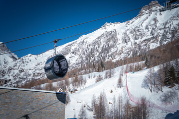 A round mirrored hot tub gondola glides at San Domenico, Italian Alps. Groomed runs, a red safety net, stone lodges, and evergreens sit under clear winter light. © Aerial Film Studio