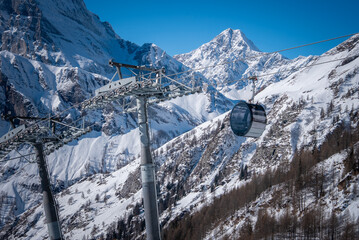 A single gondola glides over snow slopes and larch forests at San Domenico in the Italian Alps. Jagged peaks and harsh winter light cast sharp shadows, suggesting altitude. © Aerial Film Studio