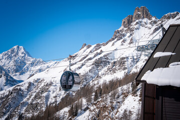 A modern gondola glides over San Domenico in the Italian Alps, beneath jagged sunlit peaks, roof snow in view, sparse larch trees on slopes, clear winter daylight. © Aerial Film Studio