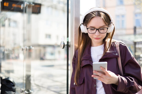 Student learning online at bus stop in city. Distance learning with smartphone. Girl with headphones outdoors. Young woman waiting for public transport, tram. Technology lifestyle, urban commute