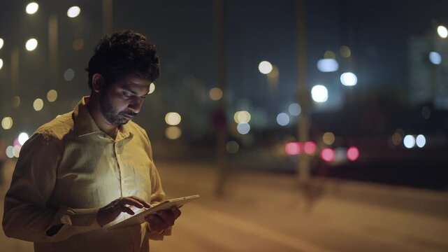 Outdoor night shot of middle-aged Indian male businessman in formal corporate office attire standing on sidewalk, busy scrolling on digital tablet, with bokeh of moving traffic and street lights
