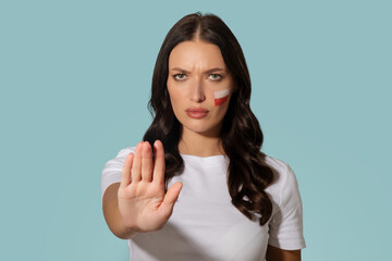 Woman with flag of Poland on cheek standing with her hand in stop gesture, posing over blue...