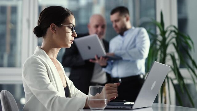 Focused woman working on laptop with business colleagues in background office. Leadership concept.
