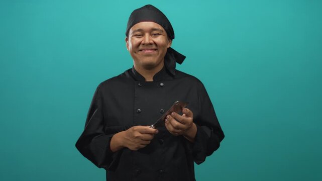 Man chef in uniform holding cleaver and smiling, showing hands in studio with teal backdrop; calm confidence.