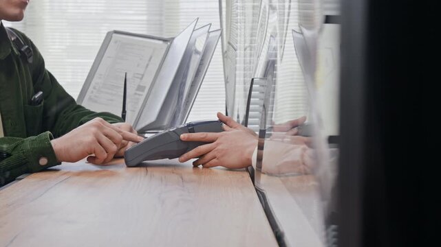 Customer entering pin on terminal, bank teller holding payment device across protective glass barrier on wooden counter