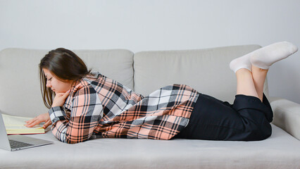 Fototapeta premium Young girl student lying with laptop and book on light sofa, online learning