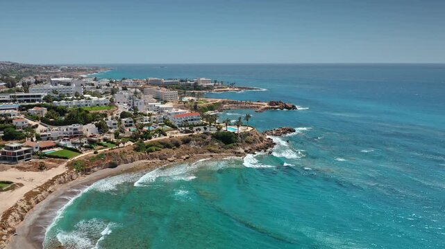 Aerial view drone flight over summer coastline of Cyprus near Paphos, showing white buildings, resorts, and a sandy beach meeting clear turquoise Mediterranean sea with gentle waves. Travel background