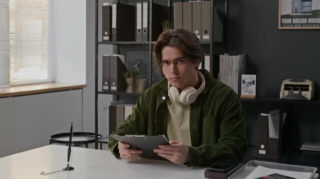 Portrait shot of young man, wearing headphones on his neck, reviewing documents at desk in bank office and looking up at camera