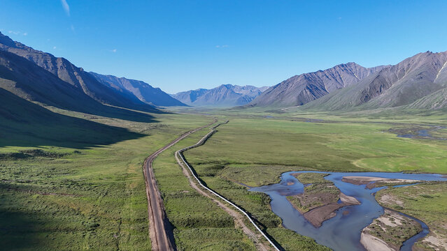Dalton highway and the trans alaska pipeline system stretching through a vast green mountain valley, with a braided river flowing under a clear blue sky near prudhoe bay, alaska, north slope