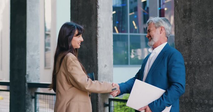 A man and a woman having a face-to-face conversation outdoors near a modern office building, shaking hands after a professional meeting and continuing their workday
