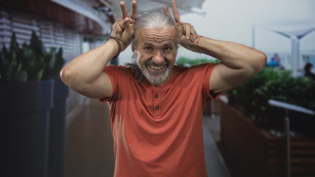 Man with long grey hair and beard makes rabbit ears with fingers behind his head at airport terminal; playful mischief.