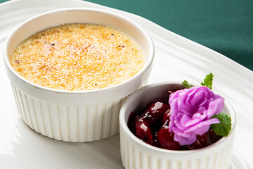 Creme brulee in a white ramekin with a side of cherry compote, garnished with a purple flower and mint leaf on a white plate and green background © pavel siamionov