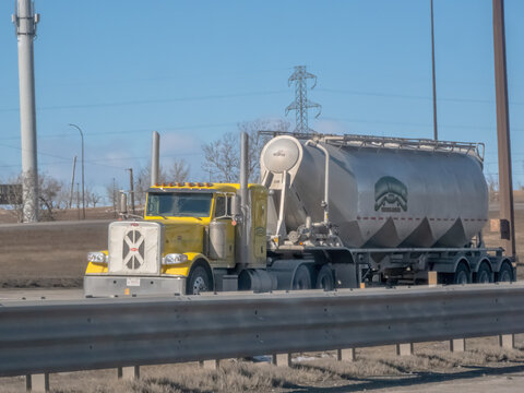 Calgary, Alberta, Canada. March 17, 2026. A yellow Peterbilt semi-truck hauls a silver bulk cement trailer along a sunny highway.