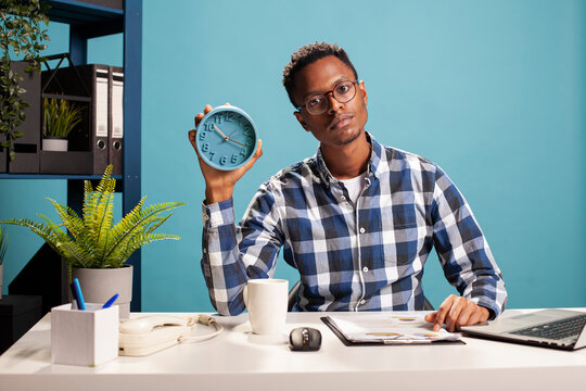 Portrait of african american freelancer sitting with a clipboard at desk and holding an alarm clock. Young business analyst handling paperwork and presenting the time with an analogue clock in hand.