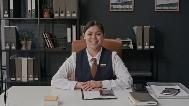 Portrait shot of young and professional bank worker looking up at camera and smiling while sitting at her office desk