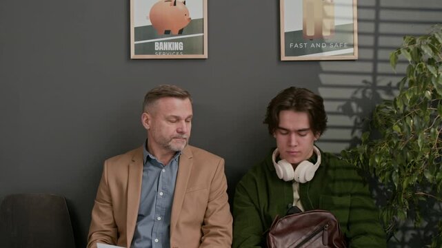 Tilt down shot of middle aged and young men sitting together in waiting area of modern bank
