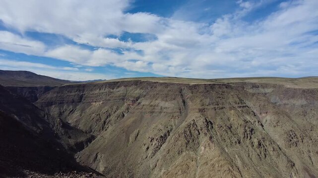 ramatic desert canyon and deep valley landscape under blue sky with clouds. Wide scenic view of Mojave desert mountain range and rugged geological formations.